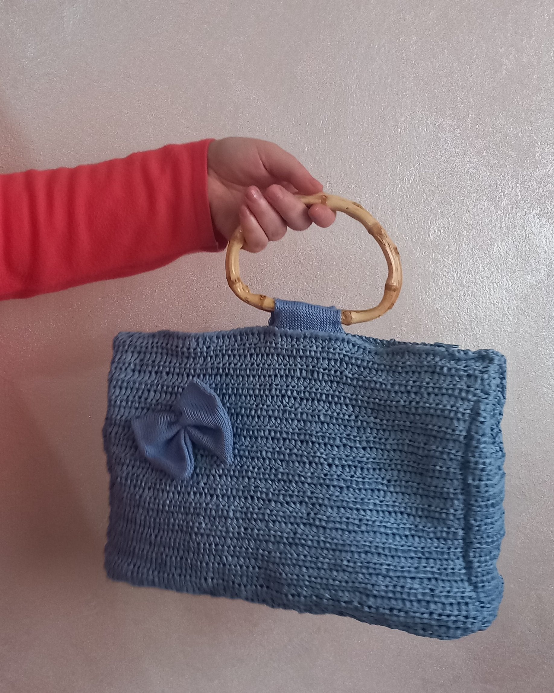 Una borsa a mano artigianale lavorata all'uncinetto in un delicato azzurro polvere. Caratterizzata da manici in bambù naturale e un romantico fiocco in tessuto tono su tono sul davanti, tenuta in mano da una persona che indossa una maglia rossa.