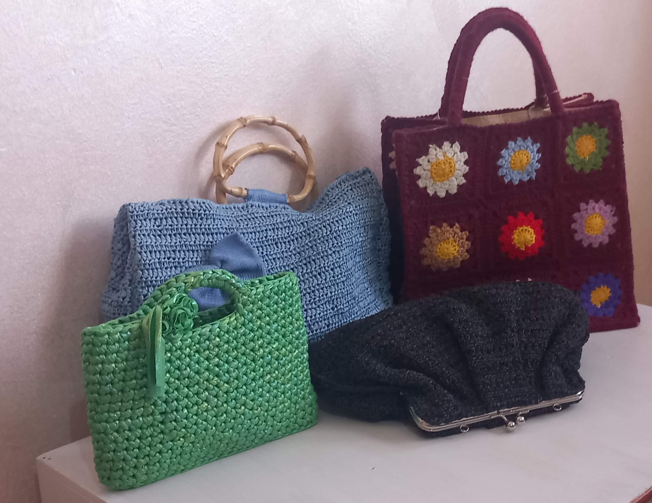 Su di un tavolo bianco sono appoggiate quattro borse all'uncinetto. Partendo a sinistra una borsa verde con un fiore (mini bag perla), dietro una borsa in rafia con un fiocco in cotone e i manici in bambu (borsa romantica). Affiancata a questa borsa c'è una in misto lana composta da diverse biatrelle floreali. Ogni fioreha un colore diverso su base bordeux. Distesa davanti a queste due una borsa nera con chiuura a pressione.