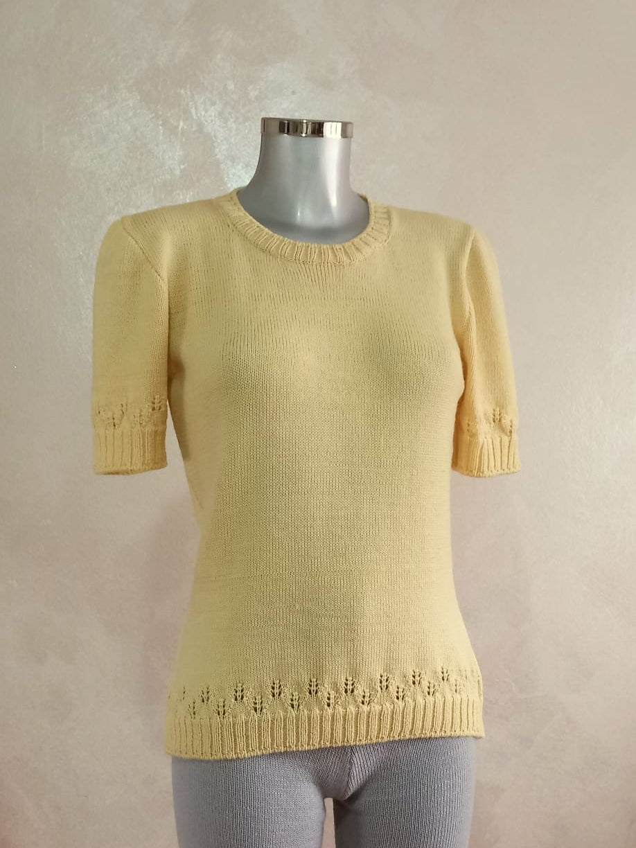 Un manichino che veste una maglia color giallo burro. La maglia ha le maniche corte e un motivo traforato che richiama le spighe di grano. Questo motivo compare sui bordi della maglia (maniche e fondo).