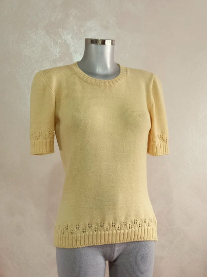 Un manichino che veste una maglia color giallo burro. La maglia ha le maniche corte e un motivo traforato che richiama le spighe di grano. Questo motivo compare sui bordi della maglia (maniche e fondo).