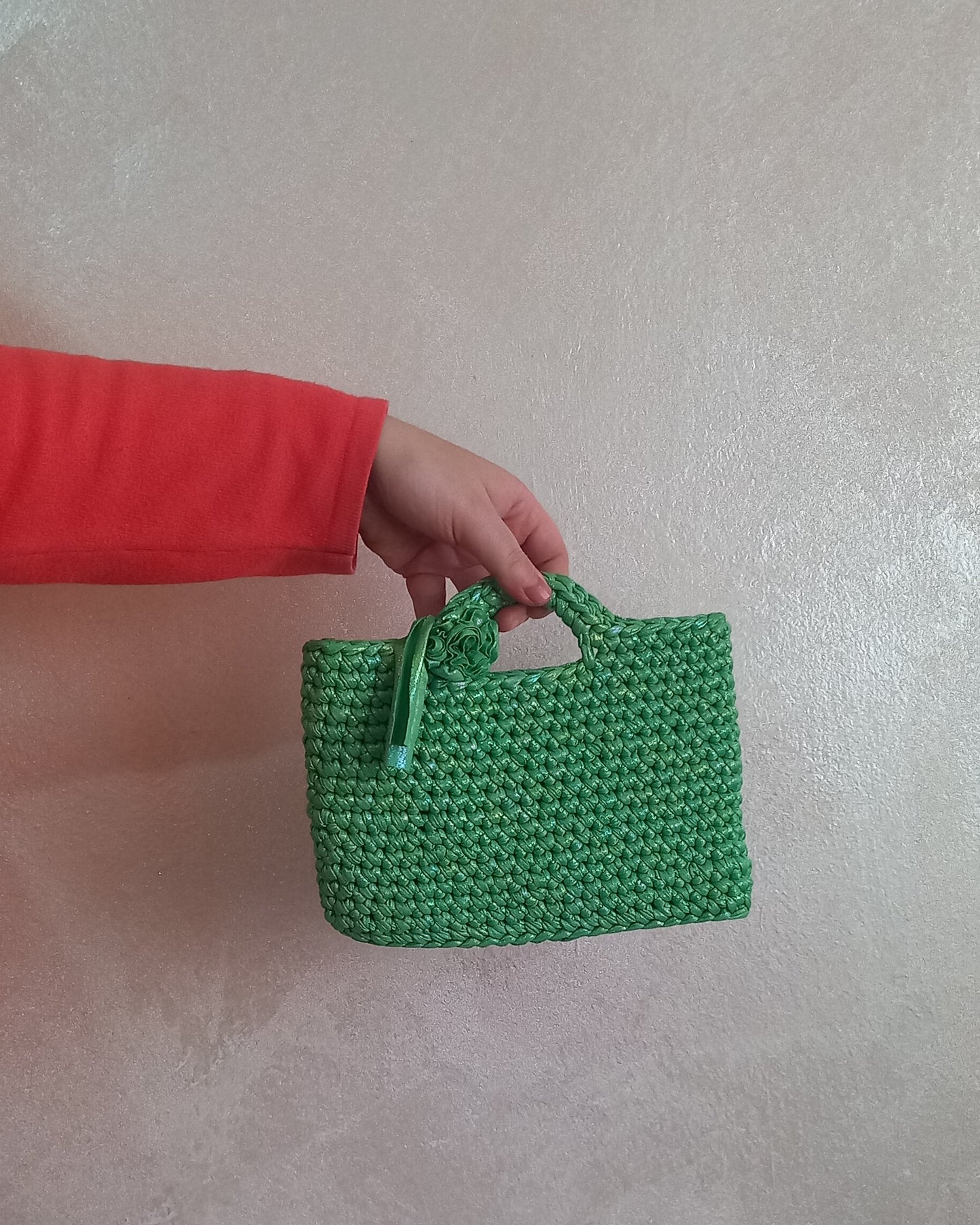 Mano che regge una piccola borsa artigianale verde irridescente. La mini bag presenta una lavorazione a maglia compatta, un manico rigido e un fiore in tessuto applicato sul lato.