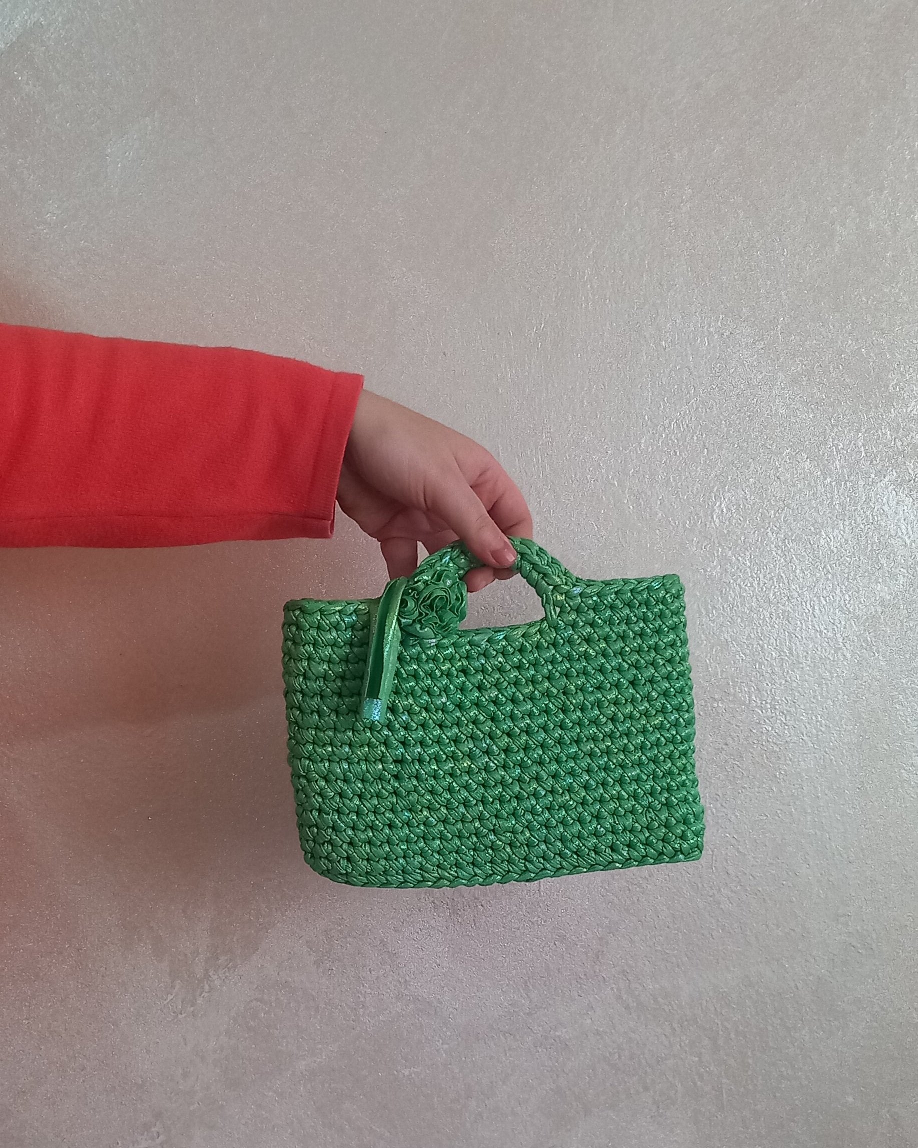 Mano che regge una piccola borsa artigianale verde irridescente. La mini bag presenta una lavorazione a maglia compatta, un manico rigido e un fiore in tessuto applicato sul lato.