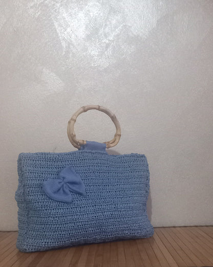 Inquadratura a tre quarti della borsa in rafia romantica. Questa borsa in raffia è color carta da zucchero con maniglie in bamboo. Impreziosita da dettagli in cotone come il fiocco.