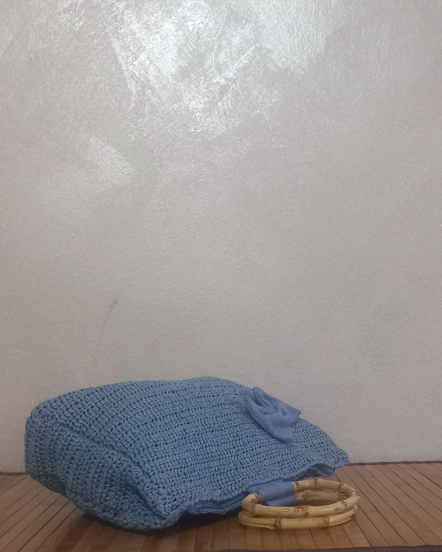 La borsa romantica all'uncinetto azzurro polvere è distesa orizzontalmente su una superficie di legno. La lavorazione a maglia morbida si piega leggermente, mostrando il lato e il fondo della borsa. Un manico in bambù naturale è visibile appoggiato sul legno, mentre un lembo di tessuto azzurro coordinato e una piccola decorazione a fiocco sono sulla parte anteriore. Lo sfondo è una parete neutra e leggermente strutturata.