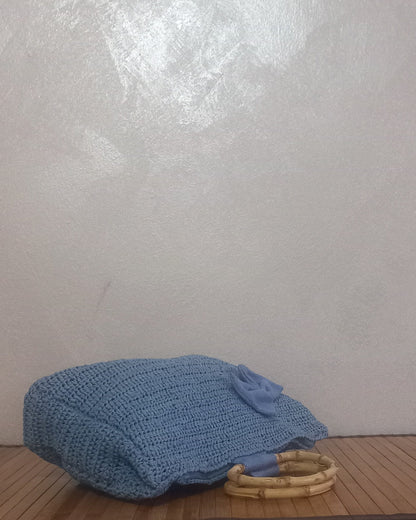 La borsa romantica all'uncinetto azzurro polvere è distesa orizzontalmente su una superficie di legno. La lavorazione a maglia morbida si piega leggermente, mostrando il lato e il fondo della borsa. Un manico in bambù naturale è visibile appoggiato sul legno, mentre un lembo di tessuto azzurro coordinato e una piccola decorazione a fiocco sono sulla parte anteriore. Lo sfondo è una parete neutra e leggermente strutturata.