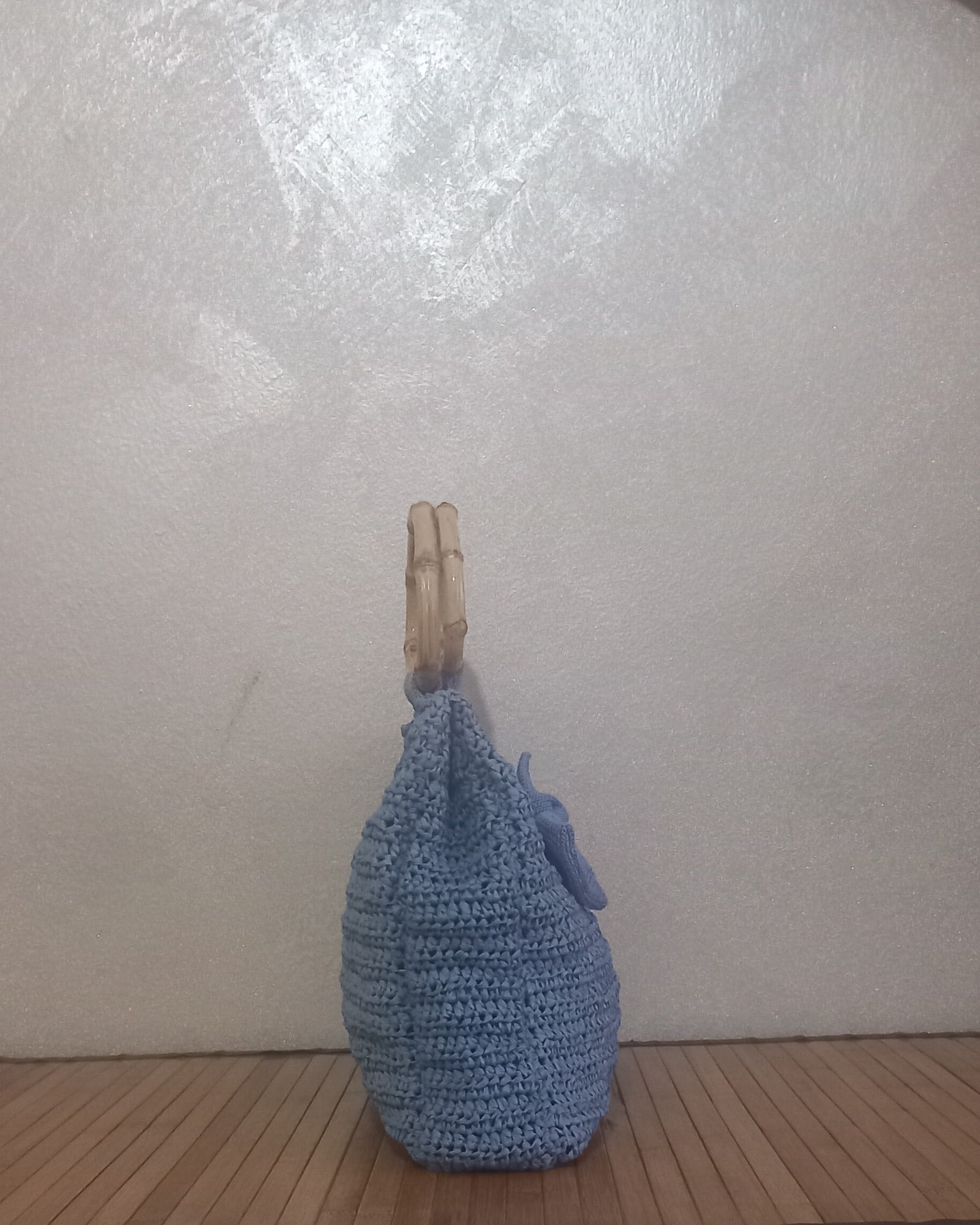 Una inquadratura laterale che evidenzia la profondità e la struttura della borsa artigianale azzurro polvere. La lavorazione all'uncinetto forma delle coste orizzontali definite. Si vedono chiaramente il profilo sottile del manico rigido in bambù e la base appoggiata sui listelli di legno. La piccola rosellina all'uncinetto è parzialmente visibile sul lato.