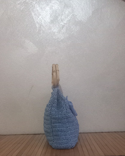 Una inquadratura laterale che evidenzia la profondità e la struttura della borsa artigianale azzurro polvere. La lavorazione all'uncinetto forma delle coste orizzontali definite. Si vedono chiaramente il profilo sottile del manico rigido in bambù e la base appoggiata sui listelli di legno. La piccola rosellina all'uncinetto è parzialmente visibile sul lato.