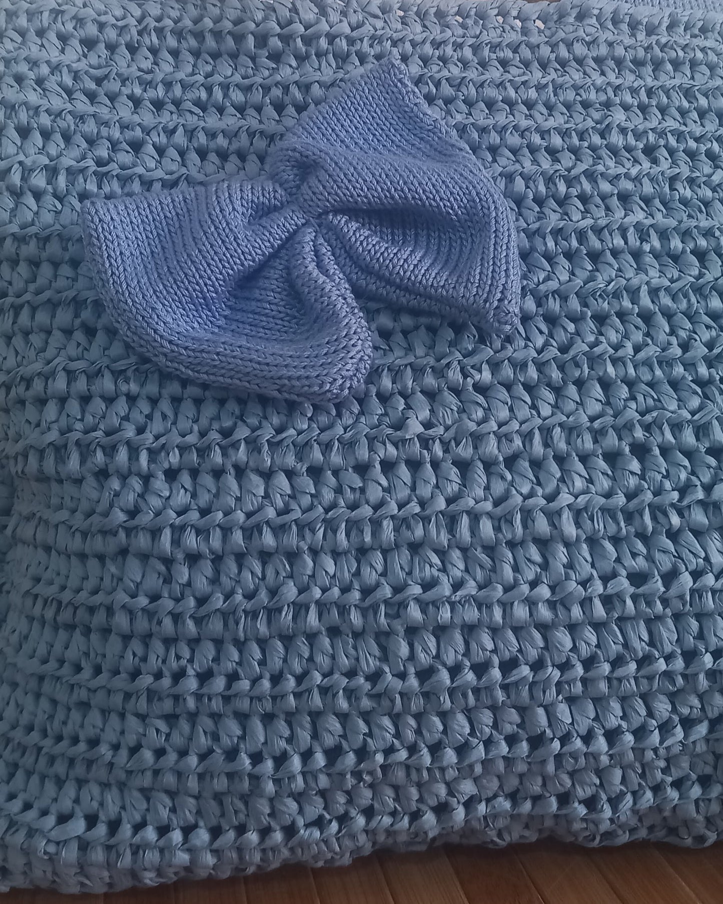 Primo piano sul particolare del fiocco della borsa romantica: un'elegante applicazione in filato azzurro polvere lavorata a maglia rasata. La sua forma morbida contrasta con la trama più densa e strutturata della borsa sottostante, anch'essa all'uncinetto in azzurro. Al centro del nodo del fiocco spicca un piccolo punto luce blu cobalto, aggiungendo un tocco di colore.