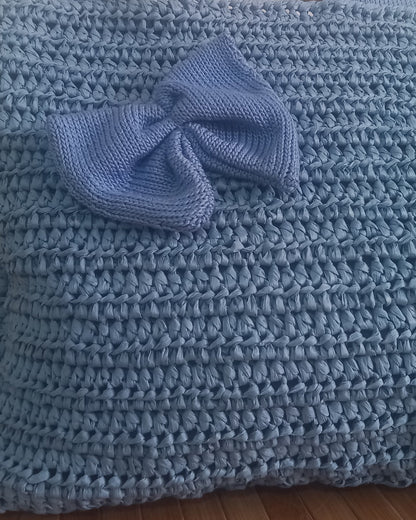 Primo piano sul particolare del fiocco della borsa romantica: un'elegante applicazione in filato azzurro polvere lavorata a maglia rasata. La sua forma morbida contrasta con la trama più densa e strutturata della borsa sottostante, anch'essa all'uncinetto in azzurro. Al centro del nodo del fiocco spicca un piccolo punto luce blu cobalto, aggiungendo un tocco di colore.