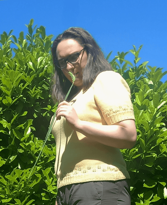 donna che tiene in mano una spiga circondata dalla natura mentre indossa una maglietta estiva in lana merino giallo burro con motivi a traforo che richiamano delle spighe
